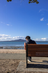 Fototapeta premium Young adult male sitting on the bench facing the sea at Mission Bay Beach, Auckland New Zealand NZ