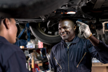 Fototapeta na wymiar Smiling black African mechanic man in overall uniform working underneath lifted vehicle in auto repair shop, Car Mechanic Concept