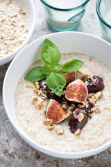 Close-up of oats porridge with fresh fig fruits, dates and nuts served in a white bowl, vertical shot