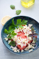 Salad with fresh watermelon, feta cheese, parsley and basil served in a blue bowl, vertical shot on a light-grey granite background