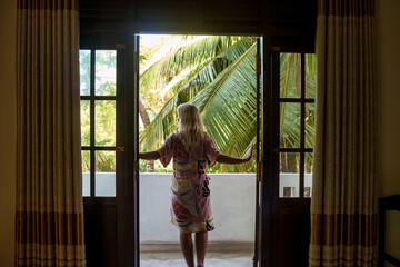 Rear view tourist young woman open door hotel balcony and goes out wearing home bathrobe in morning in countryside Sri Lanka. Vacation, travel, journey, trip and relax concept. Copy text space - obrazy, fototapety, plakaty