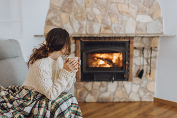 Young woman sitting at home by the fireplace with a hot tea or coffee mug and warming her hands,...