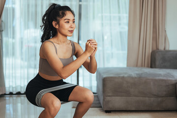 Fototapeta na wymiar Asian woman exercise at home. Young healthy female in sportswear workout doing squat with rubber band in living room, Health care and wellness concept.