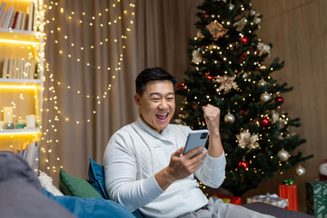 Christmas man near tree at home reading good news from phone got win and discount in online casino, Asian man holding hand up gesture of triumph sitting on sofa in living room on New Year holidays.