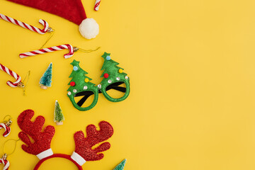 Fototapeta na wymiar Colourful funny Christmas eyeglasses, candy canes, deer horns, Santa hat, toy fir trees on yellow background. New Year, Christmas accessories for winter holidays party celebration