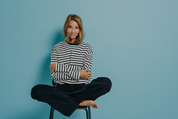 Relaxed young girl in striped shirt sitting with legs folded together in lotus pose on top of chair