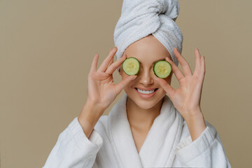 Indoor shot of pleased woman covers eyes with cucumber slices nourishes skin smiles happily dressed...