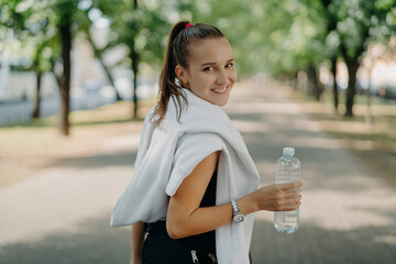 Back view of cheerful glad sporty woman with pony tail stays hydrated after cardio workout in park...