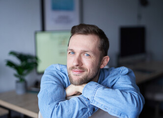 Attractive bearded male sitting in home office looking at camera smiling. Young handsome man in denim shirt, positive emotion with computer monitor on background. High quality image