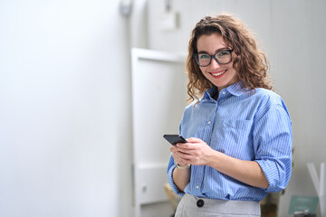 Smiling young business woman user, happy businesswoman wearing glasses holding cellular smartphone working standing in office using mobile cell phone working on cellphone looking at camera. - obrazy, fototapety, plakaty