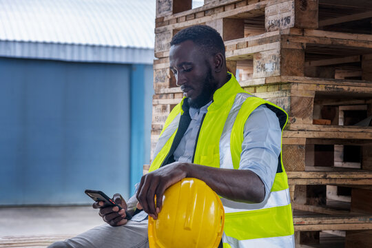 Industry Worker Sitting On Wooden Boxes In Ware House With Smart Phone On Relaxed Time After Work.