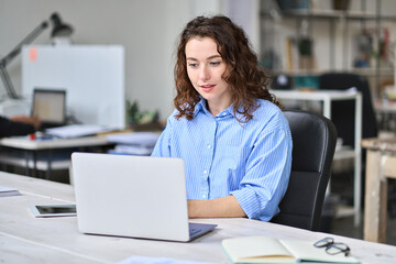 Young business woman company employee sitting at desk working on laptop. Busy female professional worker marketer using computer in corporate modern office managing data technology operations.
