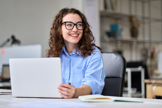 Young happy business woman company employee, marketing manager sitting at desk working on laptop. Pretty female professional worker using computer in corporate modern office looking away and smiling.