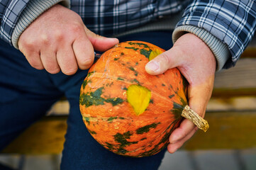 On the eve of Halloween, a man carves a spotted gourd with a small scalpel. Warm clothes outdoors on an autumn evening. Good hobby in the city park on the bench. Clise up. Selective focus