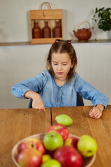 Fototapeta premium Happy little girl twist the pear, the bowl with variety fruits on table at home