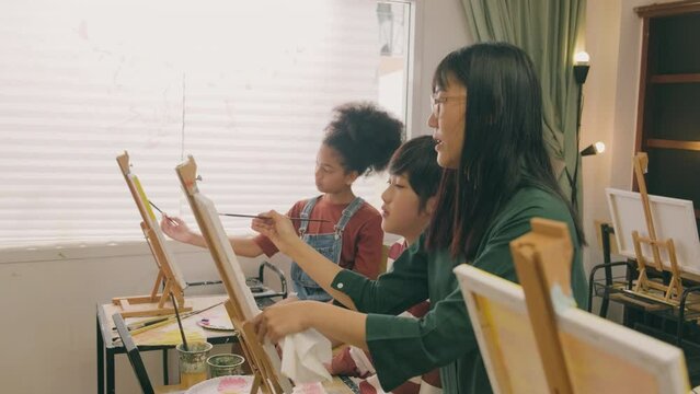 A female Asian teacher teaches and demonstrates to the children on acrylic color picture painting on canvas in art classroom, creatively learning with skill at the elementary school studio education.