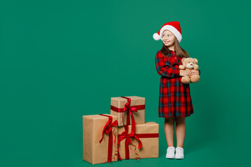 Full body merry little child kid girl 7 year old wears red dress Christmas hat posing near present box gift hold teddy bear look aside isolated on plain green background Happy New Year 2023 concept.