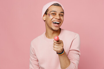 Young happy fun cheerful satisfied cool smiling gay man wear sweatshirt hat eating giving lollipop isolated on plain pastel light pink color background studio portrait. Lifestyle lgbtq pride concept.