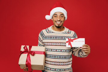 Merry young man in Christmas sweater Santa hat posing hold store gift certificate coupon voucher...
