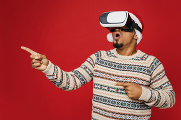 Merry young man wear Christmas sweater Santa hat posing watch in vr headset pc gadget point index finger aside on area isolated on plain red background Happy New Year 2023 celebration holiday concept