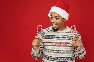 Merry surprised shocked fun cheerful cool young man wear warm cozy Christmas sweater Santa hat posing hold candy cane isolated on plain red background. Happy New Year 2023 celebration holiday concept.