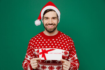 Merry young man 20s in red warm knitted sweater Santa hat posing hold gift certificate coupon...