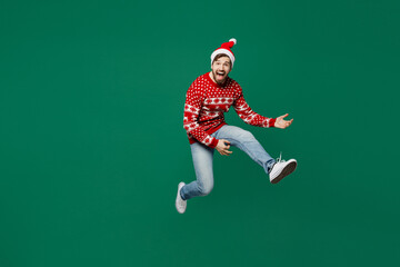 Full body merry young man wear red knitted sweater Santa hat posing jump high do hand gesture pov play guitar isolated on plain dark green background studio. New Year 2023 holiday celebration concept.