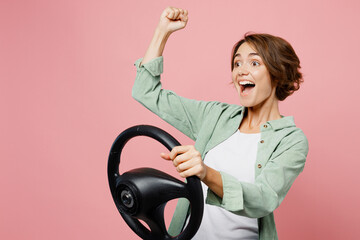 Young shocked surprised woman 20s she wear green shirt white t-shirt hold steering wheel driving car do winner gesture isolated on plain pastel light pink background studio People lifestyle concept.