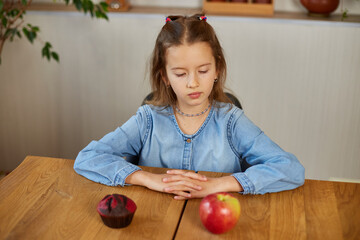 Fototapeta premium Little girl looking on sweet cake and fresh red apple and choosing in kitchen