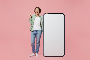 Full body young proactive woman wear green shirt white t-shirt near big huge blank screen mobile cell phone smartphone with mockup area point finger up isolated on plain pastel light pink background.