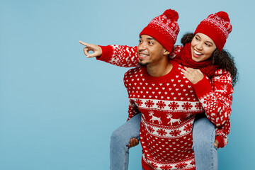 Merry young couple man woman wear red Christmas sweater Santa hat posing giving piggyback ride to...
