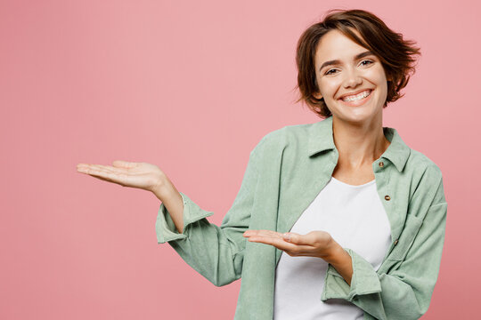 Young smiling cheerful happy cool woman 20s wear green shirt white t-shirt point hands arms aside on workspace area mock up copy space isolated on plain pastel light pink background studio portrait.
