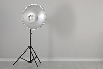 Professional beauty dish reflector on tripod near grey wall in room, space for text. Photography...