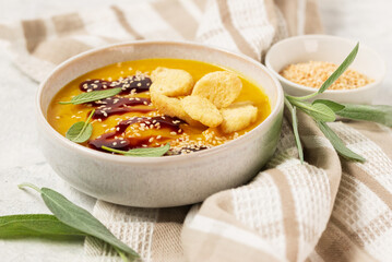 Pumpkin soup puree with sauce, sage leaves, cheese and sesame seeds in a white bowl with breadcrumbs on a light table