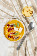 Pumpkin soup puree with sauce, sage leaves, cheese and sesame seeds in a white bowl with breadcrumbs on a light table, top view
