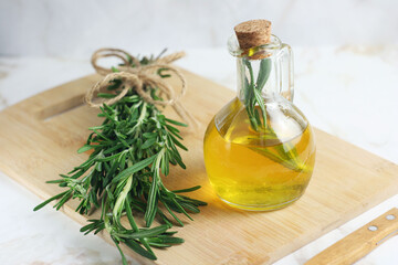 fresh rosemary lies on a wooden board on the table. Nearby is olive oil in a bottle. There is a knife nearby.