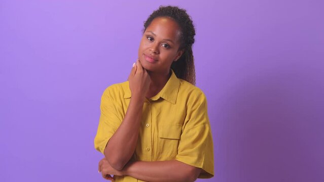 Young attractive ethnic African American woman with curly hair touches chin and looks modestly at camera dressed in casual clothes stands on plain purple background. Natural beauty concept