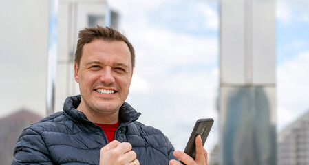 Millennial male look at camera while using mobile phone, celebrate success and give victory fist, standing outdoors at autumn