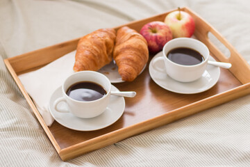 Tray of cups of coffee, croissants and apple on bed. Close-up shot of yummy and delicious romantic breakfast in bed for two in background of white and gray sheet. Food, morning concept