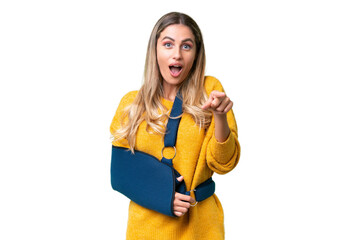 Young Uruguayan woman with broken arm and wearing a sling over isolated background surprised and pointing front