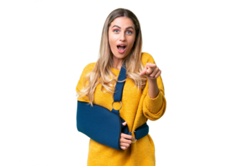 Young Uruguayan woman with broken arm and wearing a sling over isolated background surprised and pointing front