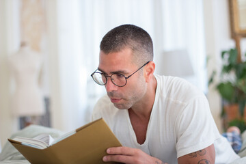 Focused man lying on bed and reading book in morning. Medium shot of concentrated Caucasian man in eyeglasses and pajamas devoting time to hobby on weekend. Morning, education concept