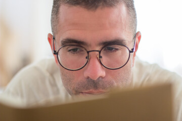 Pensive man lying and reading book in morning. Close-up shot of cheerful and handsome Caucasian man in eyeglasses devoting time to hobby on weekend. Morning, education concept