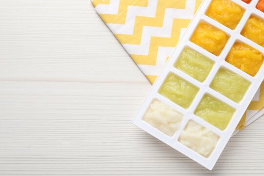 Different purees in ice cube tray ready for freezing on white wooden table, top view. Space for text