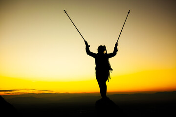 Fototapeta na wymiar Silhouette of a hiker girl on a rock pedestal with hands up. Beautiful orange sunset.