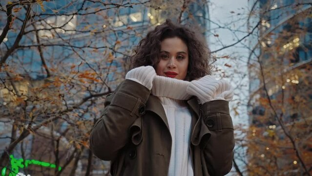 Portrait confident young curly woman touching knitted sweater neck posing in autumn city park near skyscrapers in downtown street looking at camera seductively, attractive female, urban style