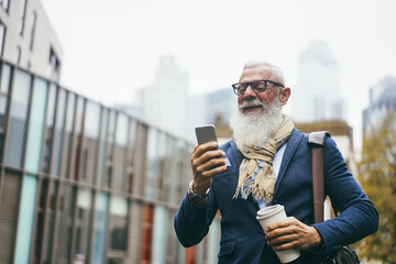 Business hipster senior man using mobile phone while walking to work with city in background - Focus face - obrazy, fototapety, plakaty