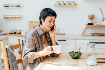 A young woman talking on the phone and drinking coffee sits at home in the kitchen.