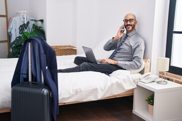 Young hispanic man business worker using laptop talking on smartphone at hotel room