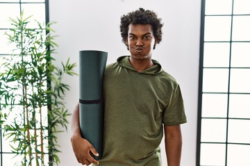 African man with curly hair holding yoga mat at studio puffing cheeks with funny face. mouth...